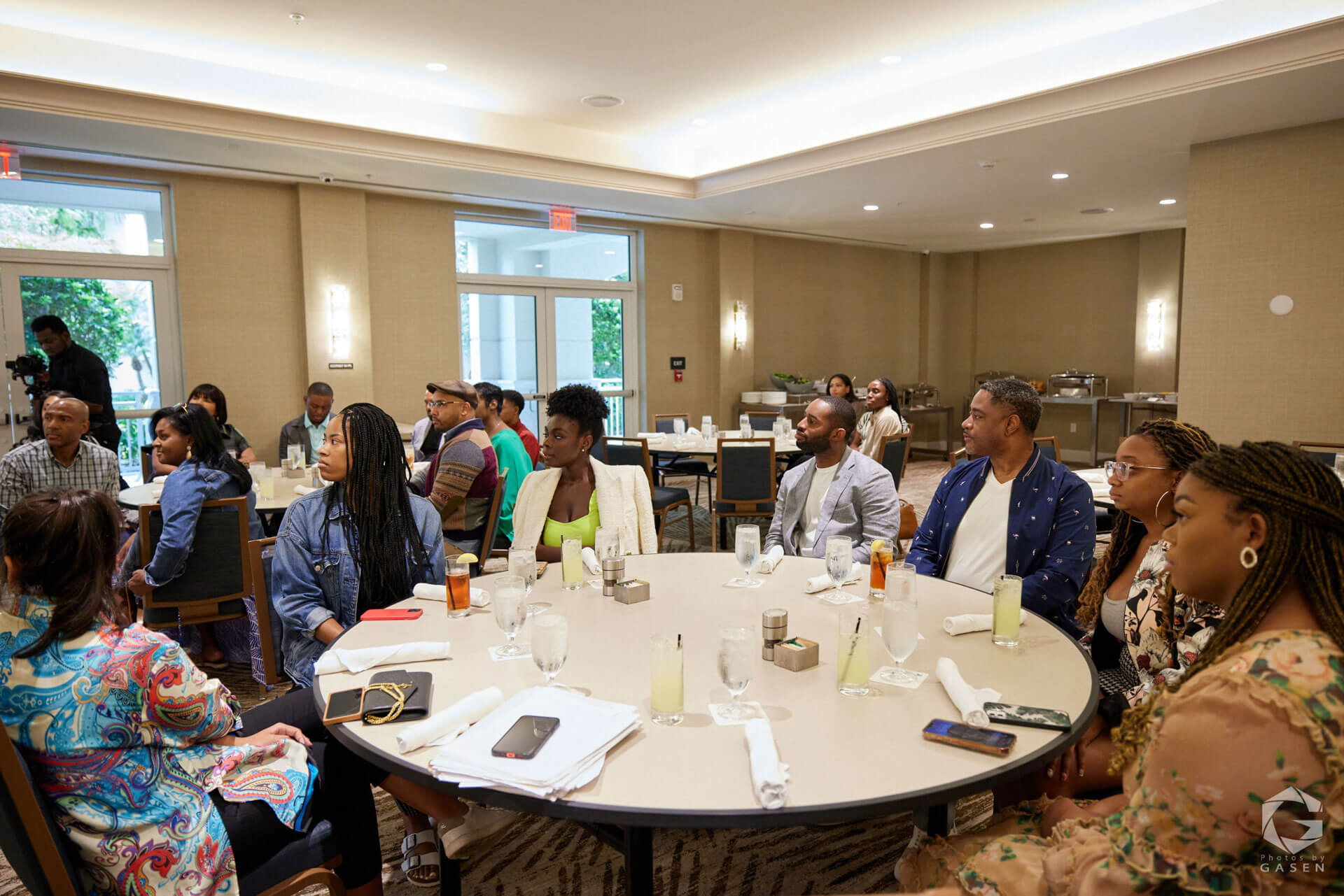 Attendees of a formal event seated around tables, attentively listening to a speaker, with a well-lit room in the background.