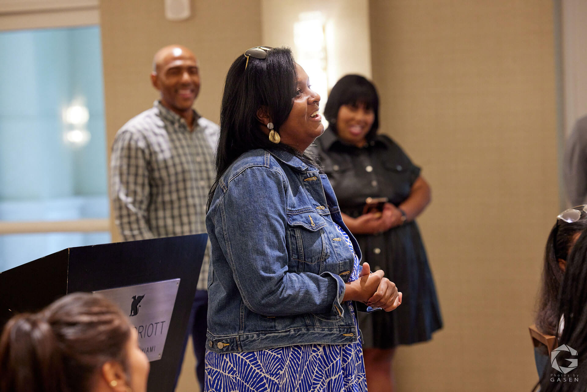 A woman in a denim jacket standing and smiling during a discussion at a formal indoor event, with people in the background.