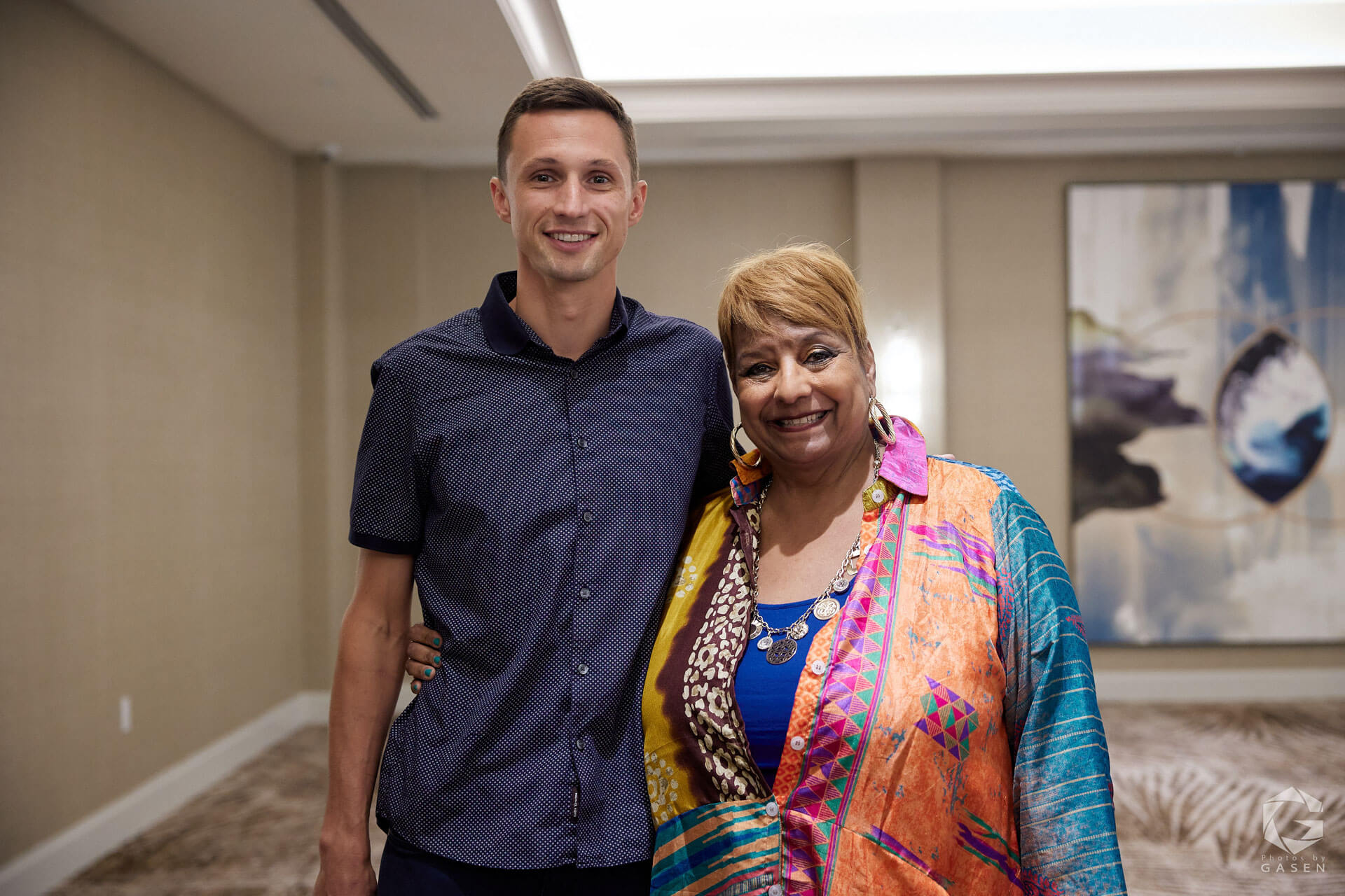 A man and a woman standing side by side, smiling at the camera in a bright and welcoming event setting.