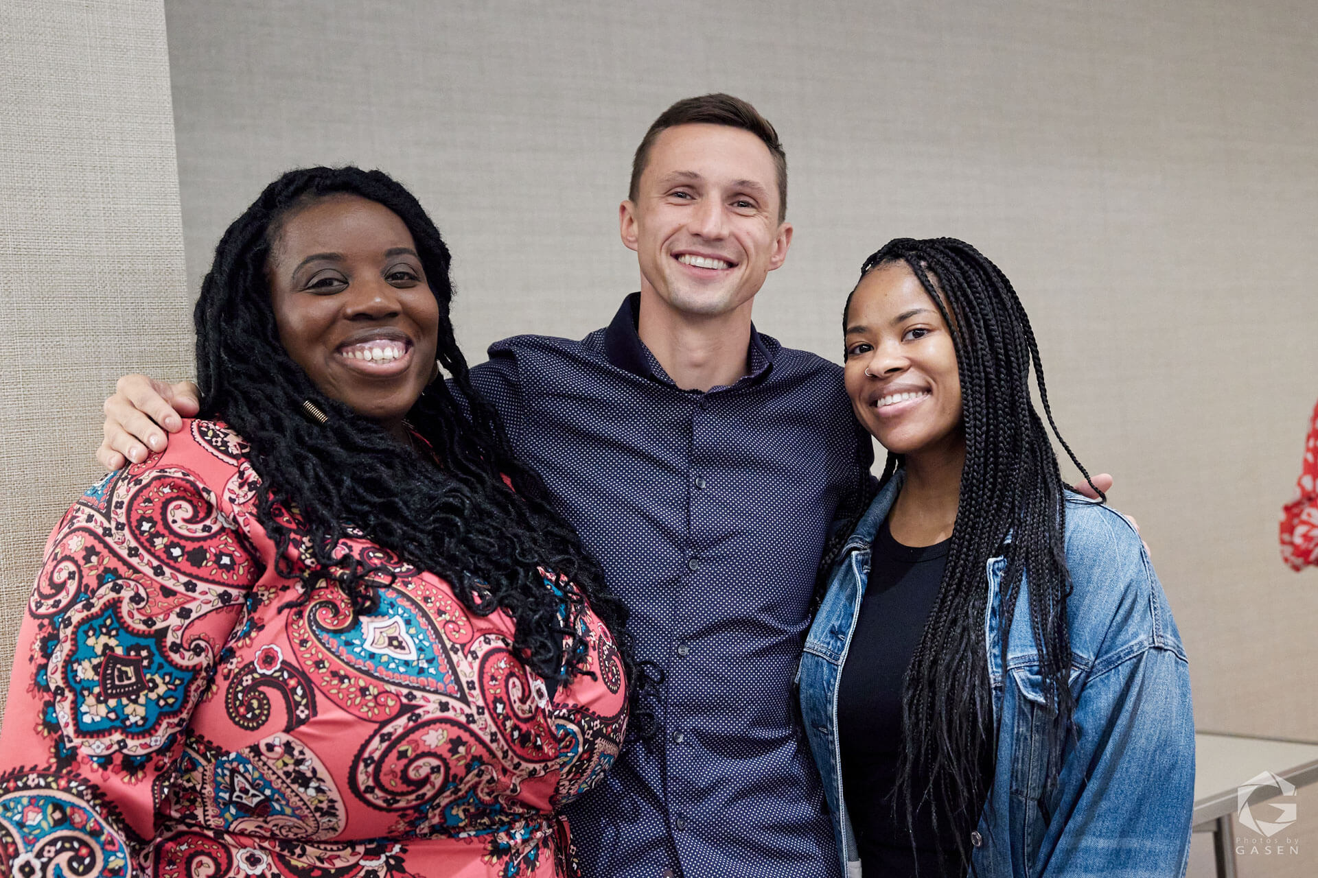 Three individuals standing side by side, smiling warmly, in a casual and welcoming setting.