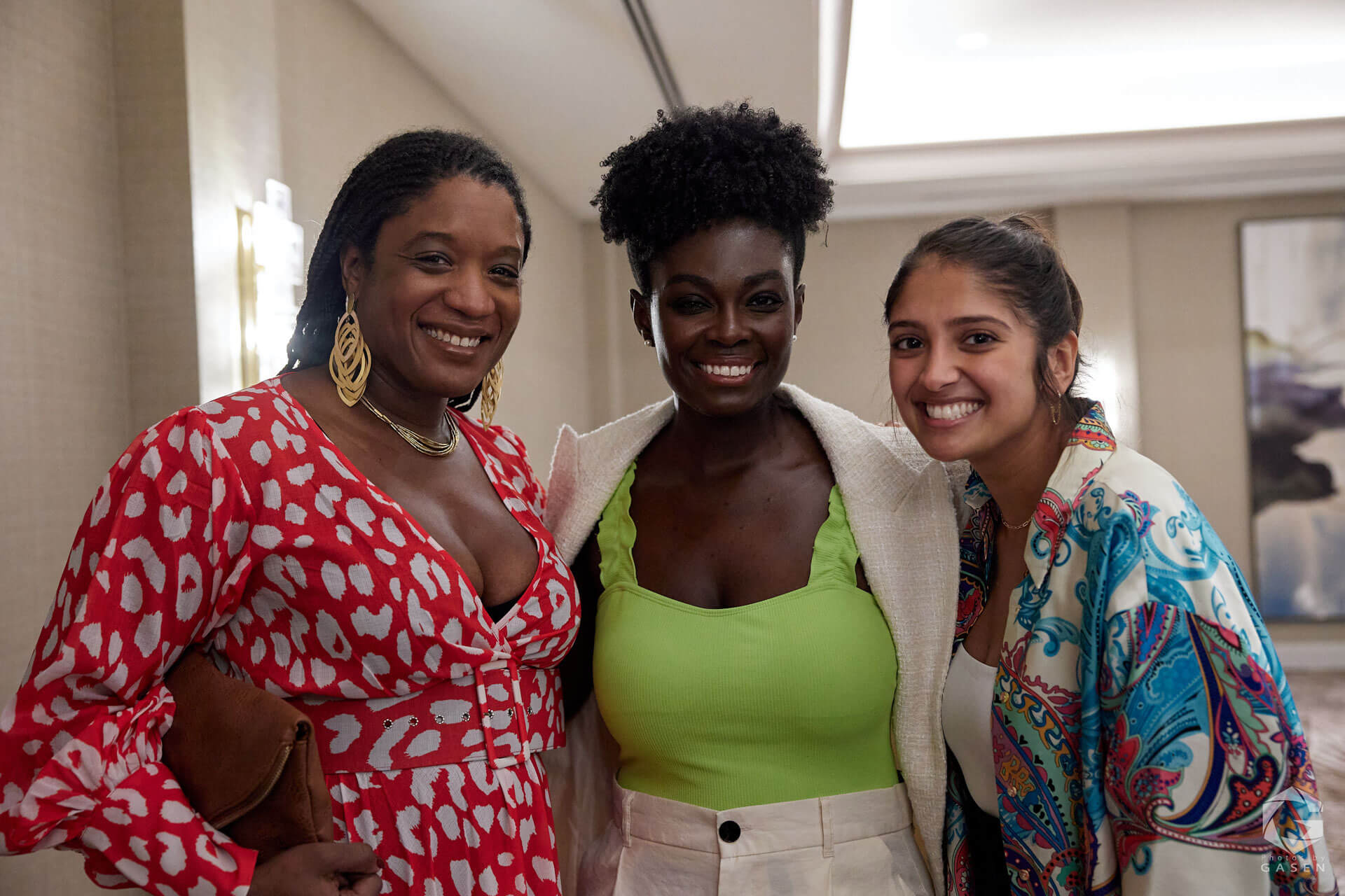 Three women standing together, smiling brightly, dressed in vibrant and colorful attire at a professional event.