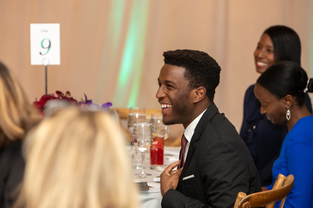 A young man in a suit and tie is seated at a round event table, laughing and engaging in conversation. He is surrounded by other attendees, some of whom are smiling. A table sign with the number "9" is visible in the background along with elegant event decor.