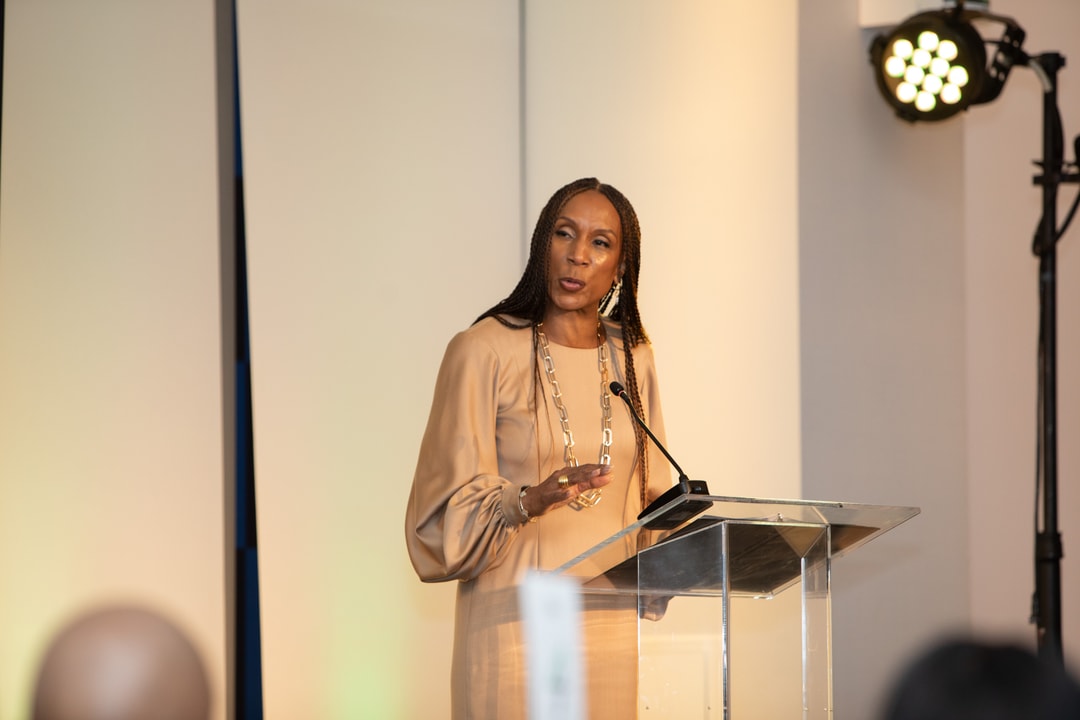 A woman in a beige dress with long sleeves and gold jewelry stands behind a clear podium, delivering a speech. She gestures while speaking, with a focused expression. The background includes professional lighting and a stage setup.