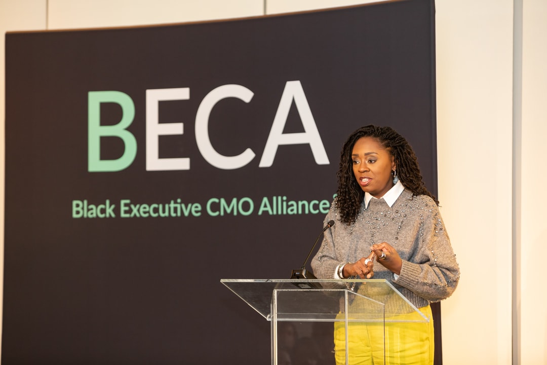 A woman in a stylish gray sweater adorned with embellishments, a white collared shirt, and bright yellow pants stands behind a podium, speaking at a BECA event. The backdrop prominently displays "BECA" and "Black Executive CMO Alliance," indicating the event’s branding.