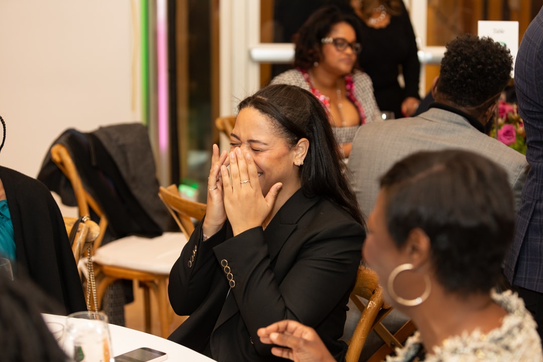 A woman in a black blazer sits at a table, covering her face with her hands while laughing. Other guests around her are engaged in conversation, and a floral centerpiece is visible in the background.