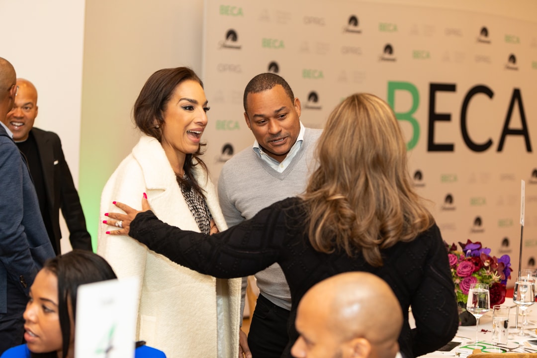 A woman in a white coat smiles while talking to another woman dressed in black at a BECA event. A man in a light gray sweater stands beside her, listening to the conversation. The background features a "BECA" branded backdrop and other guests engaged in discussions.