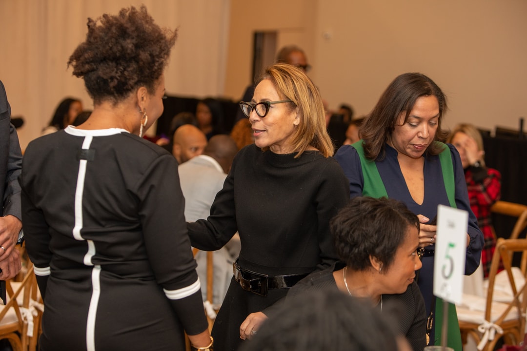A woman in a black dress with glasses is engaged in conversation with another woman wearing a black and white dress at a formal event. Other attendees are seen in the background, mingling and networking around round tables decorated with floral centerpieces. A table sign with the number "5" is visible in the foreground.