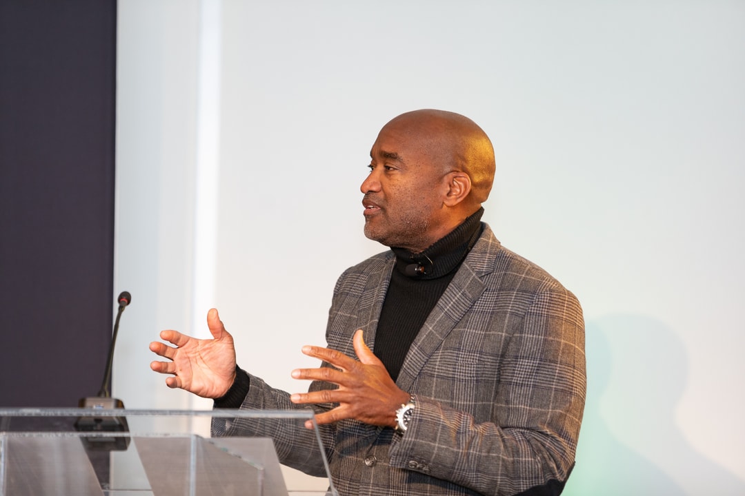 A bald man in a plaid blazer over a black turtleneck sweater stands at a clear podium, gesturing with both hands while speaking. He has a focused expression, and the background consists of a white wall and a microphone stand.