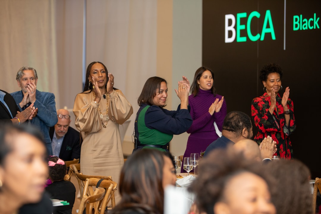 A group of attendees stands and claps during the BECA event, with some dressed in elegant attire and the large event banner visible in the background.