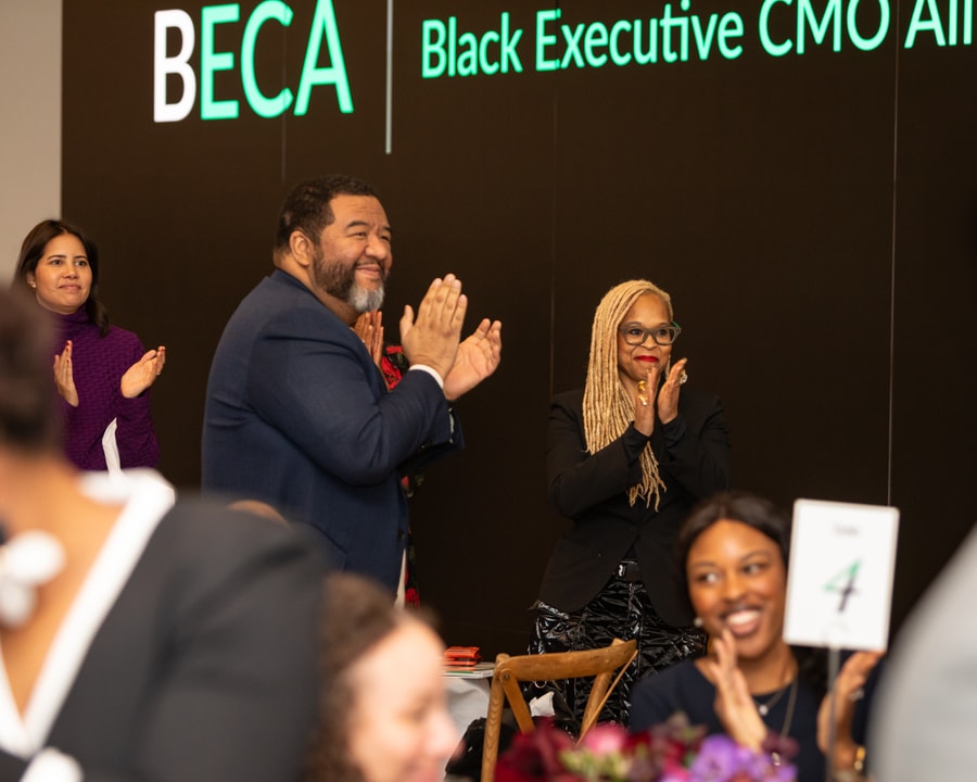 A man in a navy suit and a woman in black with long blonde braids stand clapping in front of a BECA event backdrop, while other attendees in the background also applaud.