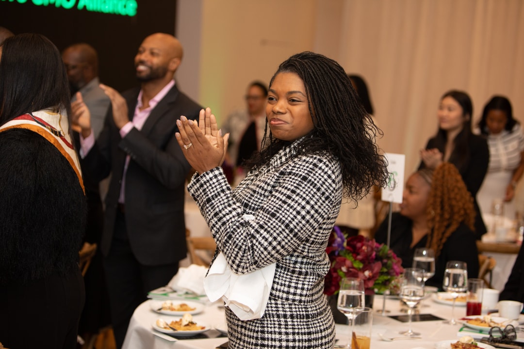 A woman in a black-and-white checked jacket stands at a table, smiling and clapping, alongside others applauding in the background at a formal event.