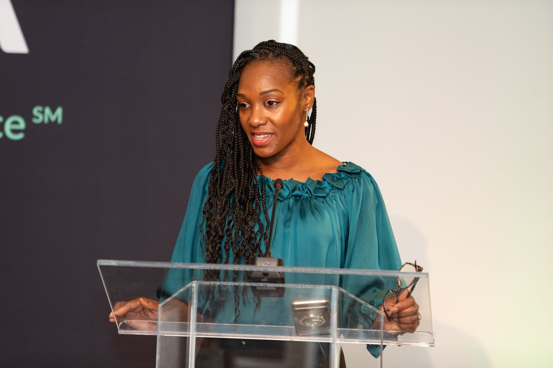 woman in a teal blouse with long braided hair stands behind a clear podium, delivering a speech. She holds a pair of glasses in one hand and speaks into a microphone, with a confident expression. A dark event backdrop is partially visible behind her.