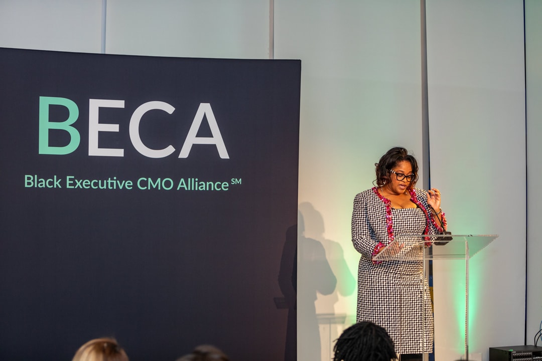 A woman in a checkered blazer with red accents speaks at a clear podium with the "BECA Black Executive CMO Alliance" banner in the background.