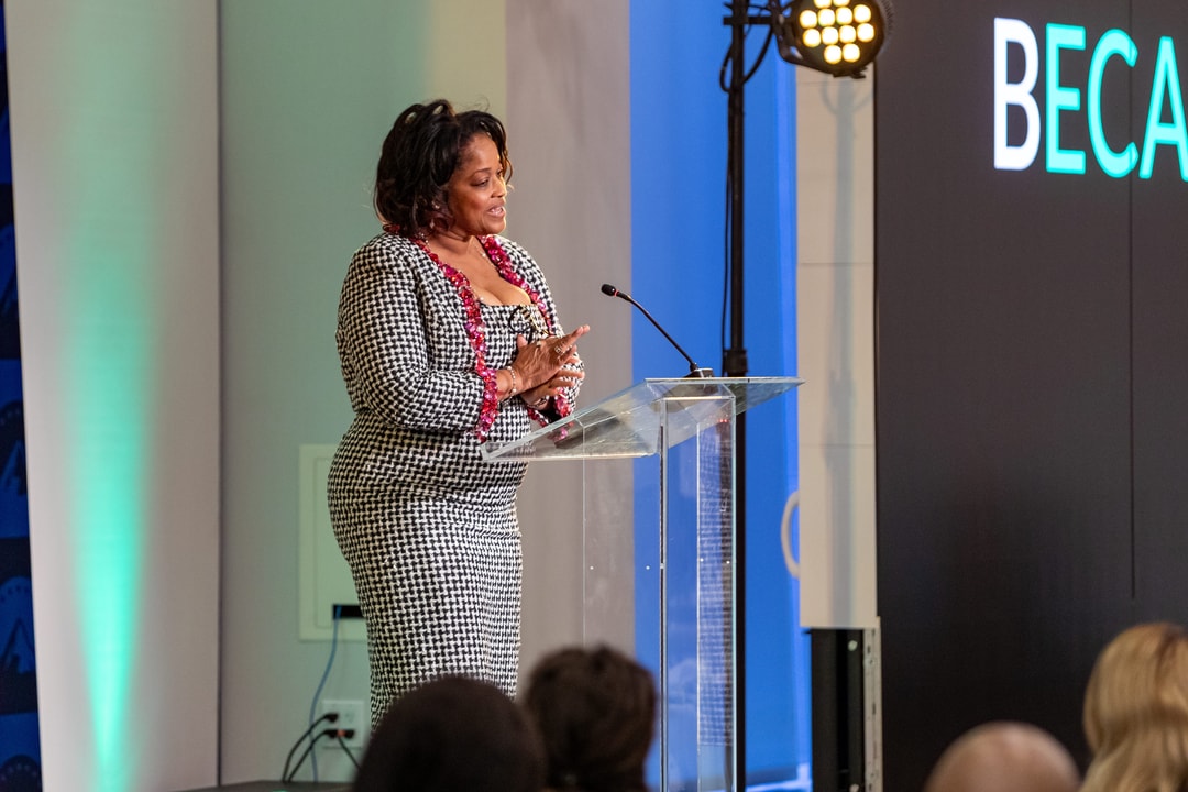 The same woman at the podium glances towards the audience with a serious expression, with bright stage lighting illuminating her.
