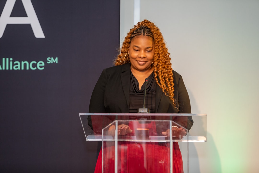 A woman with long, curly hair in a black blazer and red skirt stands behind a clear podium, looking down and smiling. She is speaking at a formal event, with a "BECA" branded backdrop behind her.