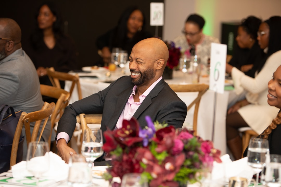 A bald man in a dark suit with a pink shirt sits at a round event table, smiling and appearing amused. The table is adorned with a floral centerpiece, and other attendees are seen in the background enjoying the event.