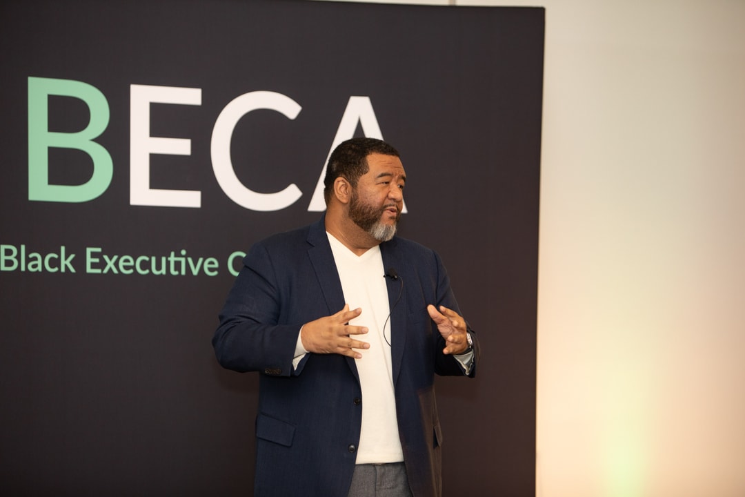 The same man is delivering a keynote speech at the BECA event, standing on stage in front of an audience. He is gesturing expressively with both hands, with the BECA logo visible on the backdrop behind him.