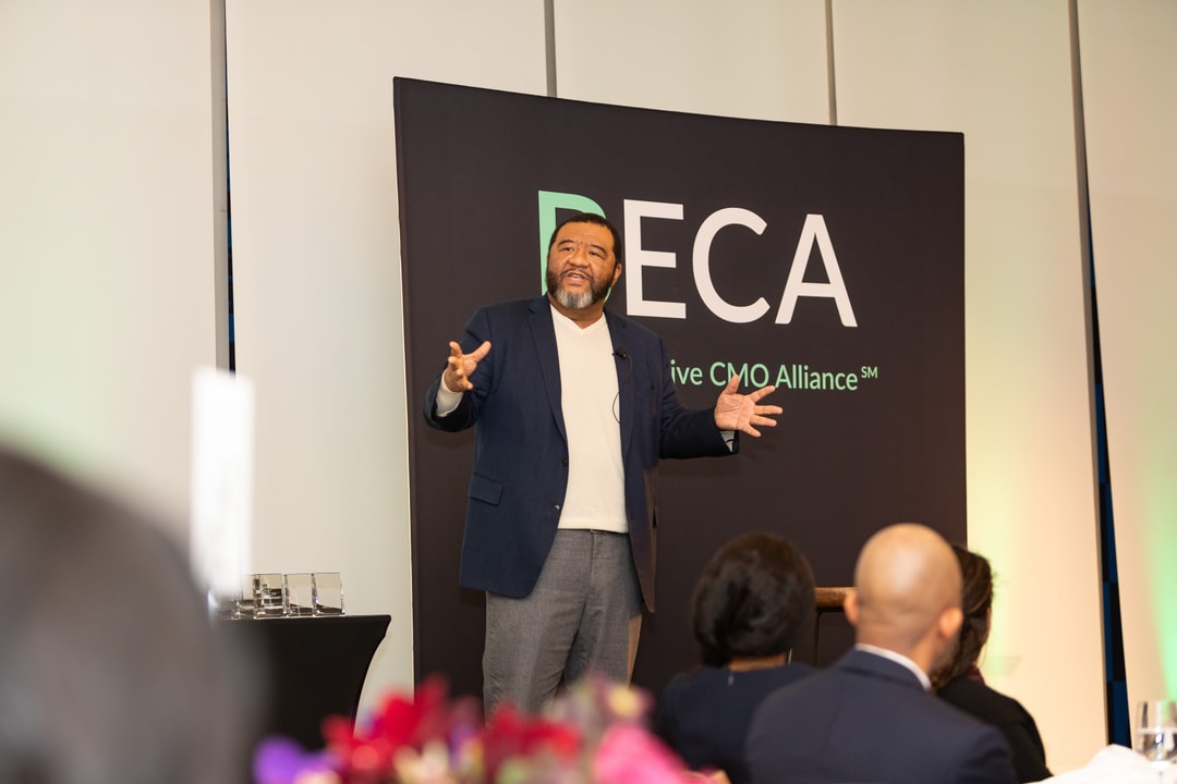 A man in a navy blazer and white shirt is speaking on stage at the BECA (Black Executive CMO Alliance) event. He is gesturing with his hands while standing in front of a black backdrop with the BECA logo and text.