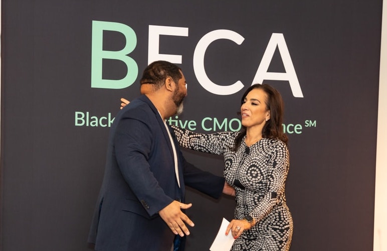 A man in a navy blue blazer and a woman in a patterned dress embrace in front of a BECA-branded backdrop, smiling warmly as they greet each other.