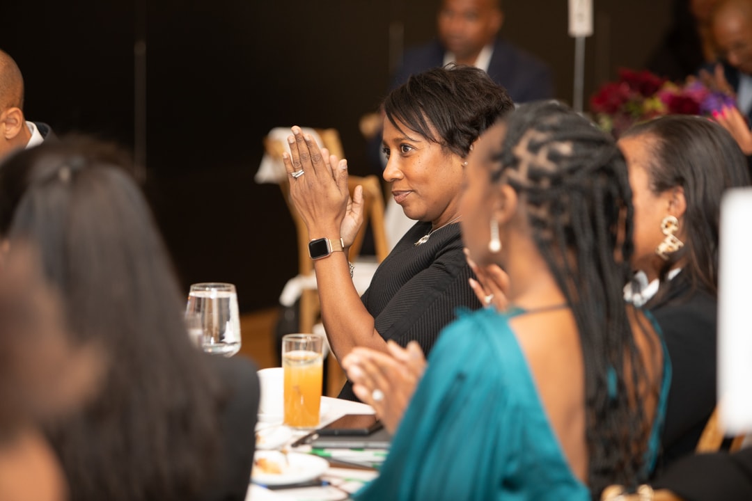 A woman in a black dress with a smartwatch claps while seated at a table, surrounded by other attendees dressed in formal wear at the BECA event.