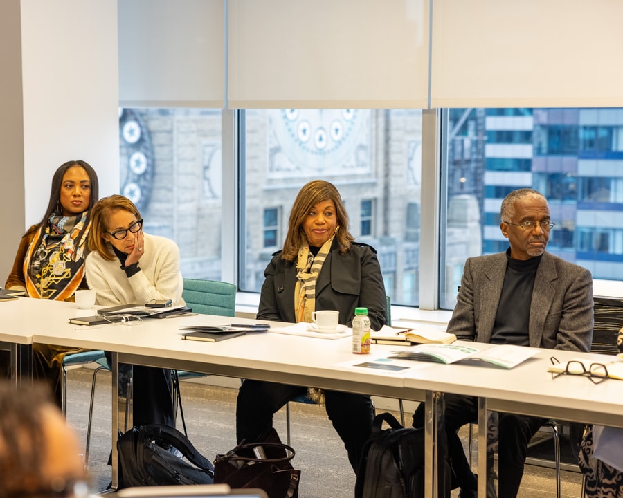 A group of professionals sit around a conference table, attentively listening and taking notes, with a large window showing a cityscape in the background.