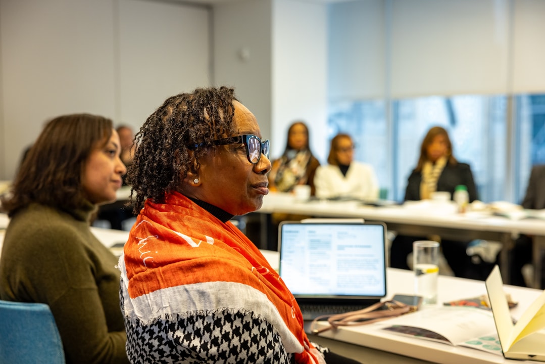 A woman wearing a red and white scarf and glasses listens attentively during a professional meeting, with a laptop open in front of her.