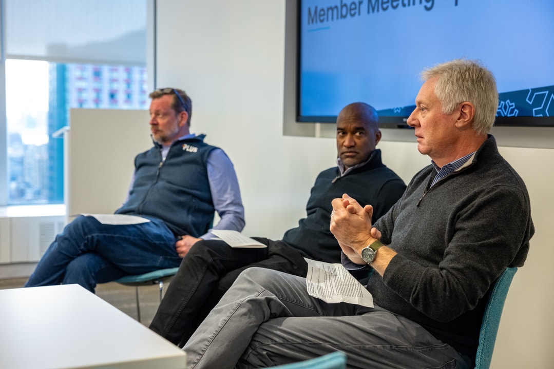 Three men sit in a panel discussion, holding papers, in front of a screen displaying "Member Meeting."