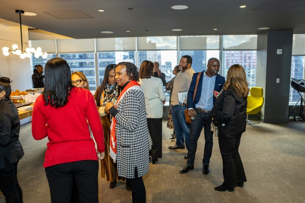 A group of professionals engages in conversation at a networking event in a bright office space with large windows showcasing a city skyline. Attendees are dressed in business casual attire, mingling and enjoying refreshments.