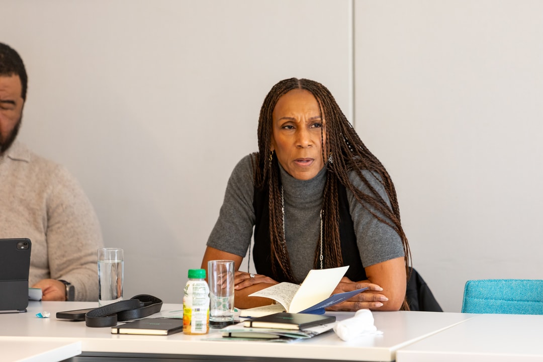 A woman with long braided hair sits at a conference table, speaking and holding a book