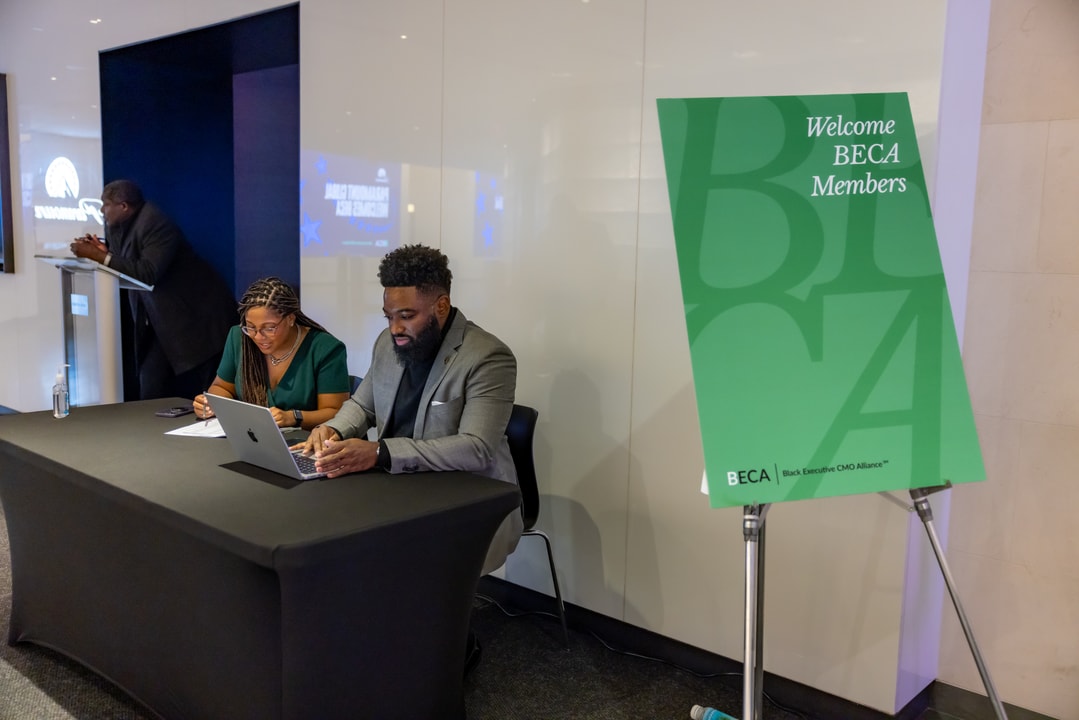 Two individuals sit at a black table working on a laptop, assisting with event check-in, while a green "Welcome BECA Members" sign stands nearby.