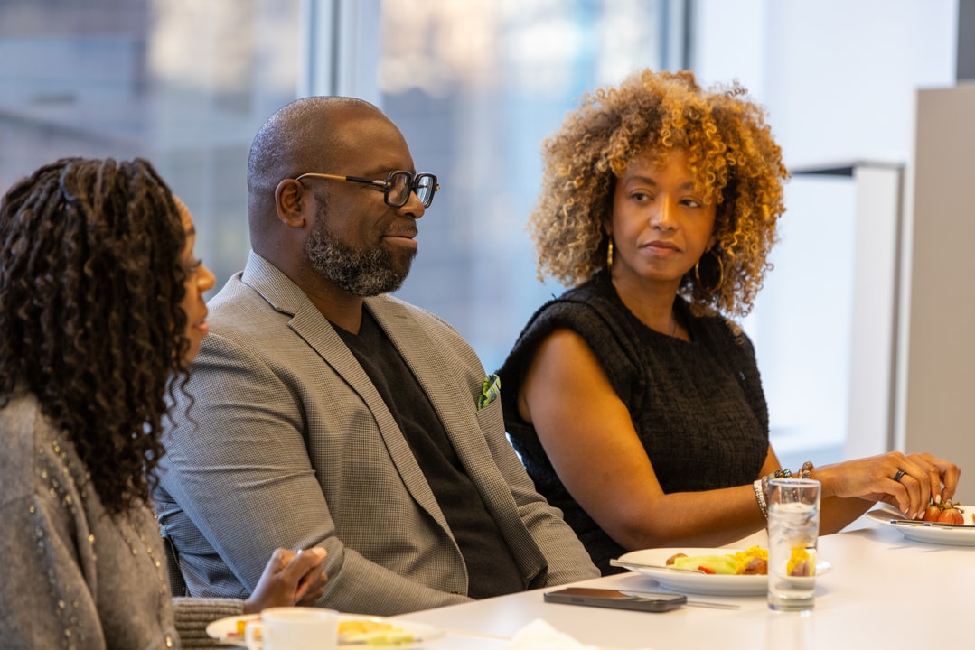 Three people sit at a table during a discussion, with one man in glasses smiling and a woman with curly blonde hair attentively listening.