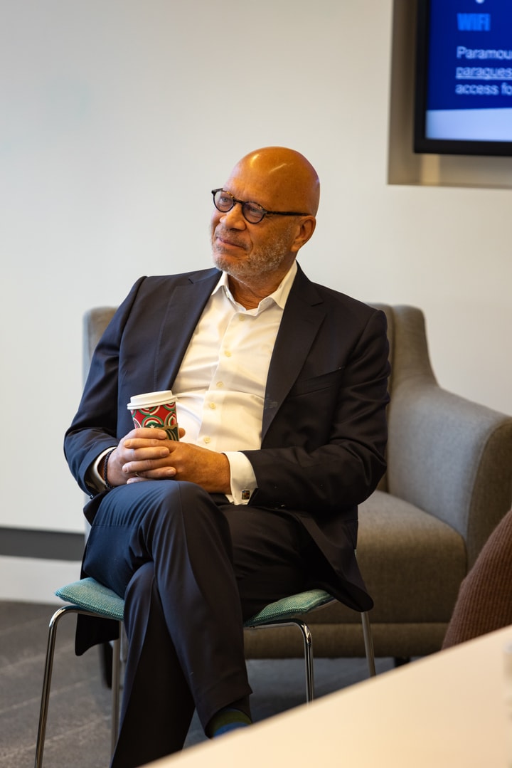 A bald man in a suit sits comfortably in a chair, holding a festive coffee cup and smiling during a discussion.