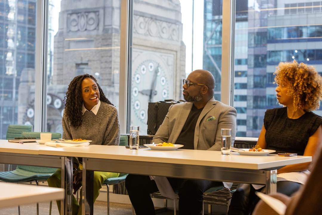 A woman with curly hair in a gray sweater is smiling while conversing with two colleagues during a meal in a modern office with large windows showing a clock tower.