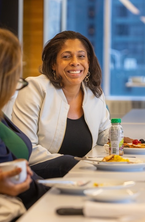 A woman in a white blazer smiles while sitting at a table, enjoying a meal with others in a well-lit conference room.