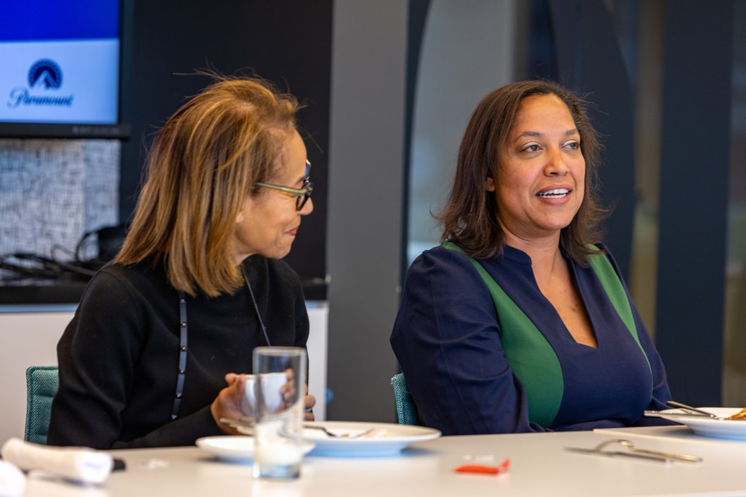 Two women at a conference table are engaged in discussion over a meal, with one actively speaking and the other attentively listening, with a Paramount logo visible on the screen in the background.