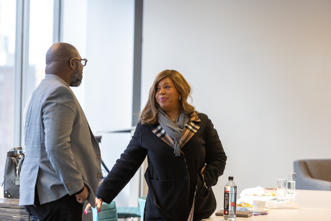 A man in a gray suit is speaking with a woman wearing a black coat and scarf. They are in a modern office environment with a conference table nearby.