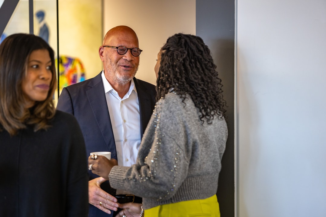 A man in glasses and a navy blue suit conversing with a woman wearing a gray sweater and yellow pants in a networking setting.