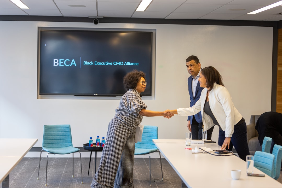 Two women shaking hands in a conference room with a BECA sign on the screen behind them. A man in a blue suit stands in the background observing.