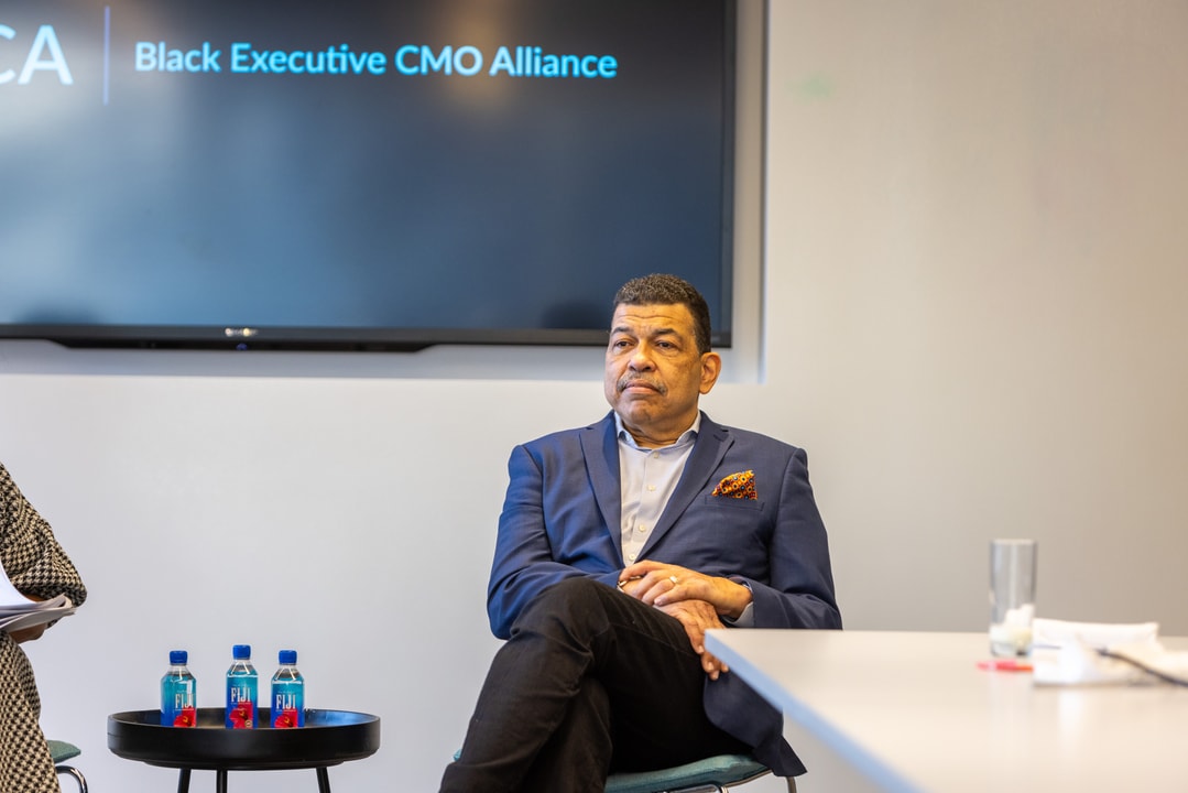 A man in a navy blue suit with an orange pocket square sits with his legs crossed in a conference room. Behind him, a screen displays "Black Executive CMO Alliance." A small table with water bottles is visible in the foreground.