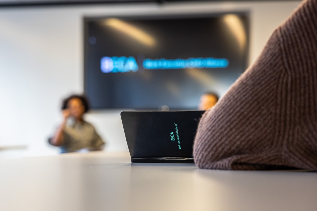 Close-up shot of a BECA-branded folder on a table, with a blurred view of a speaker and audience in the background.