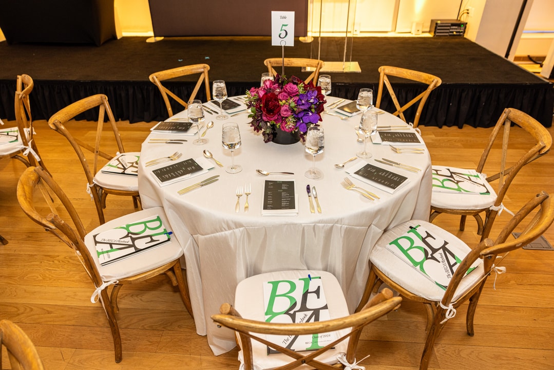 A round table at a formal event is elegantly set with white tablecloths, silverware, and glassware. A floral centerpiece with red and purple flowers sits in the middle. Each chair has a copy of The BECA Playbook placed on its seat. A table number sign displaying "5" is positioned at the center of the table. The stage is visible in the background.