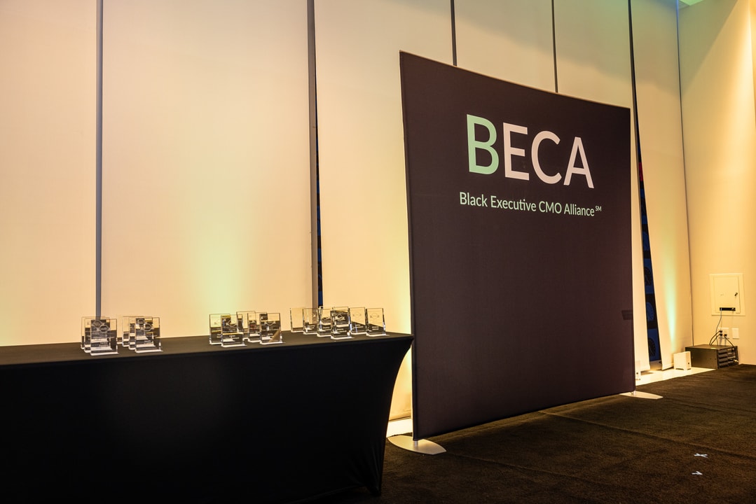 A black-draped awards table holds multiple clear glass awards, neatly arranged in rows. A large banner behind the table displays the BECA (Black Executive CMO Alliance) logo. The background features a softly lit wall, and the awards reflect the event lighting, enhancing their polished appearance.