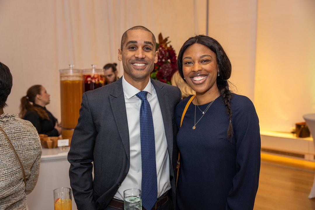 A smiling man in a navy blue suit and a woman in a navy blue dress pose together for a photo at a networking event. They stand close to each other, looking directly at the camera. Behind them, a table with large beverage dispensers and other guests mingling can be seen.