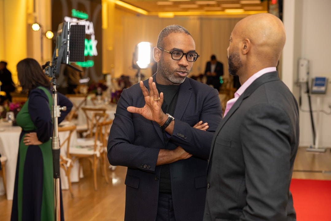 Two men in suits are engaged in a serious conversation at a formal event. One man, wearing glasses and a black blazer, gestures with his hand while speaking, while the other, in a dark suit with a pink shirt, listens attentively. The background includes event tables, floral arrangements, and professional lighting.