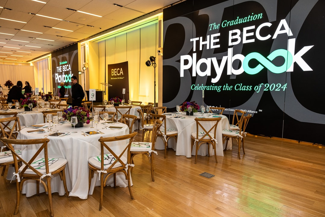 A wide-angle view of the BECA graduation event shows multiple round tables set for guests, each adorned with floral centerpieces, glassware, and copies of The BECA Playbook on the seats. A large digital screen in the background displays "The Graduation - THE BECA Playbook - Celebrating the Class of 2024." Warm lighting and professional event decor create an elegant atmosphere.