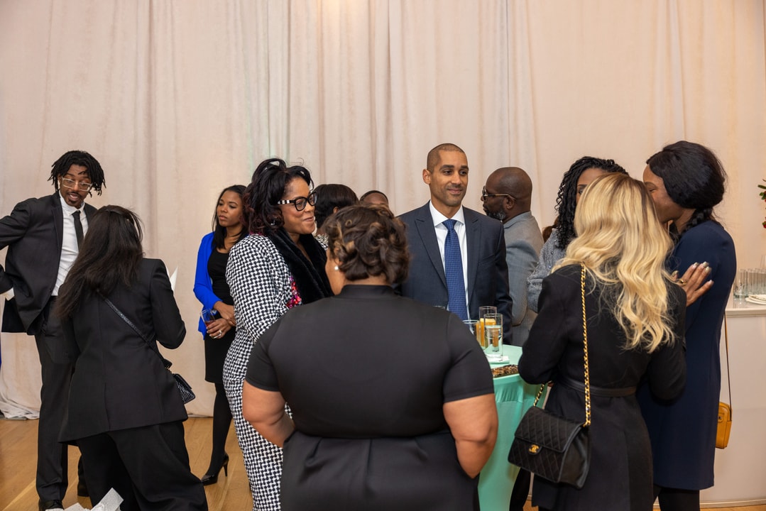 A group of people, dressed in business attire, are mingling and chatting at a professional networking event. A man in a navy blue suit and tie stands near a table with drinks, engaging with guests. Other attendees, including women wearing stylish dresses and coats, are seen conversing in small groups. The setting has soft lighting and a decorative curtain backdrop.