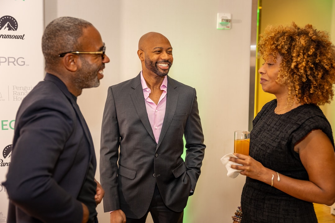 Three professionally dressed individuals are engaged in conversation at a networking event. One man in a dark suit and pink shirt is smiling while speaking to another man in glasses and a dark blazer. A woman in a black dress with curly hair holds a drink and listens attentively. The background features event signage with logos, including "Paramount" and "Penguin Random House."