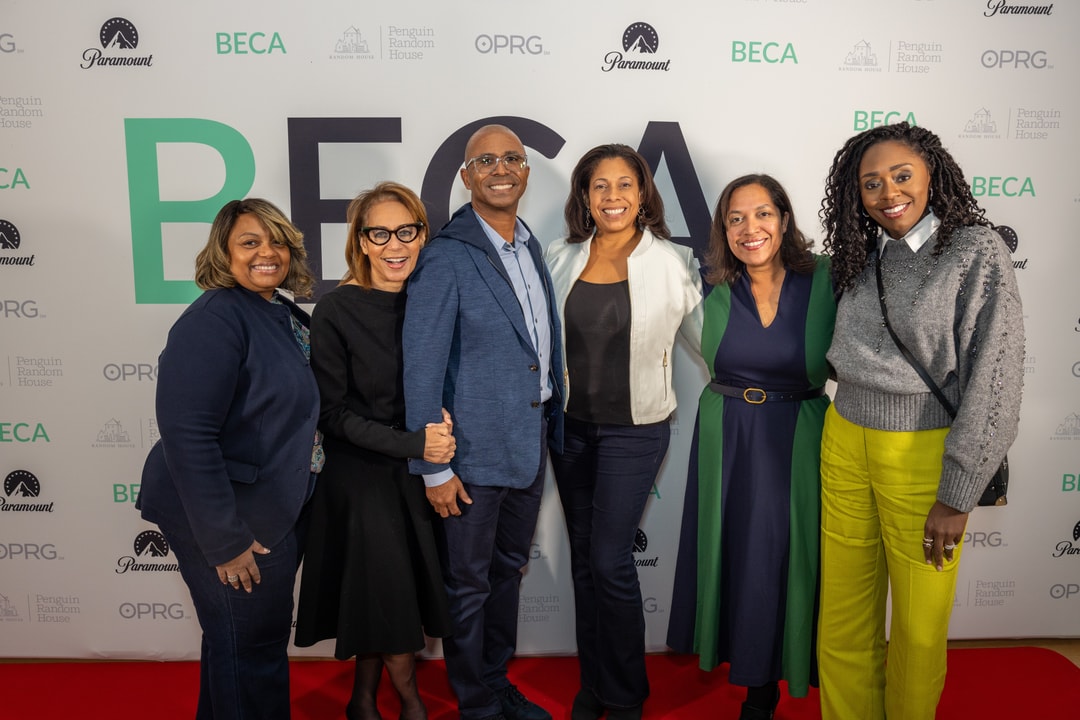 A group of six individuals pose together in front of a "BECA" event backdrop. They are dressed in a mix of professional and stylish casual attire, including blazers, dresses, and a sweater with bright yellow pants. They stand closely together, smiling warmly for the photo on a red carpeted area.