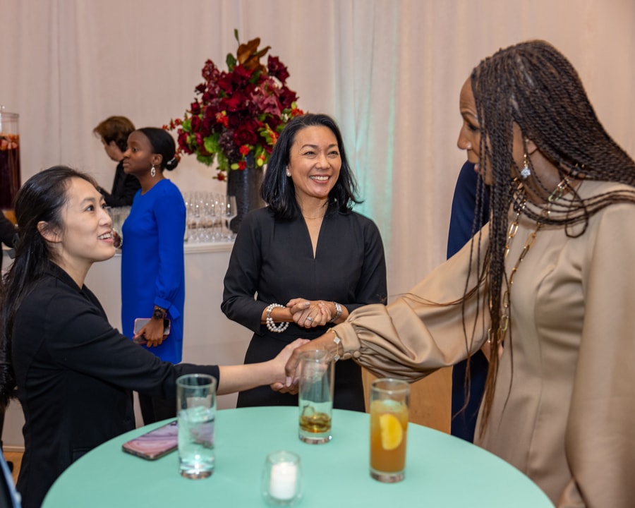 A woman in a beige dress with long sleeves and gold jewelry shakes hands with another woman seated at a round table, who is smiling. Another woman in a black dress stands nearby, smiling as she observes the interaction. The background includes a decorated table with drinks, floral arrangements, and other event attendees.