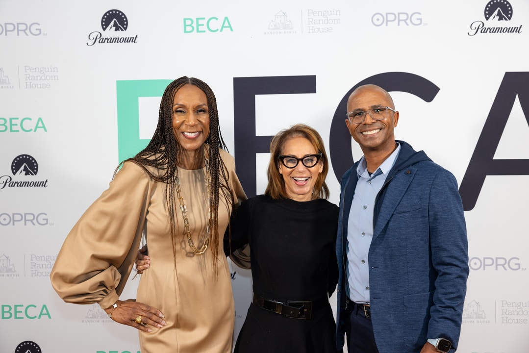 Three people stand together, smiling, in front of a sponsor-branded backdrop. The woman on the left wears a long-sleeved, tan dress and a gold necklace, the woman in the middle wears a black outfit and glasses, and the man on the right wears a blue blazer over a light-colored shirt.